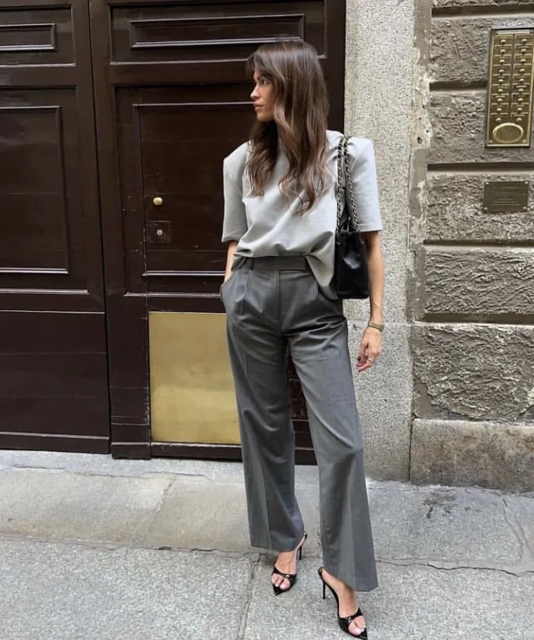 Minimalist grey trouser outfit featuring a tucked-in grey short-sleeve top, black shoulder bag, and black strappy heels, styled against a wooden door.
