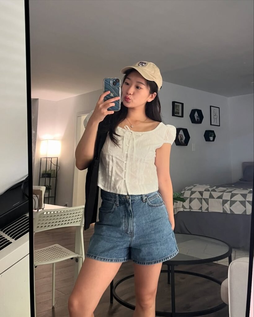 outfit korean style of a white button-up short-sleeve blouse with puff sleeves, blue high-waisted denim shorts, and a beige baseball cap.