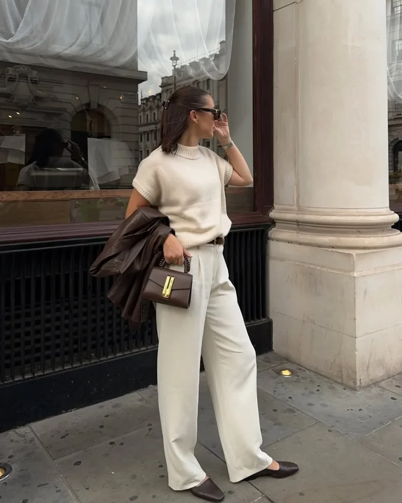 Office outfit in light neutrals – cream sleeveless knit top tucked into wide-leg cream trousers, styled with brown pointed flats, a brown leather jacket, and a matching structured handbag.
