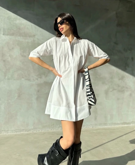 Woman wearing casual summer dress in plain white shirt-dress style with short sleeves, accessorized with black knee-high boots and a striped bag.