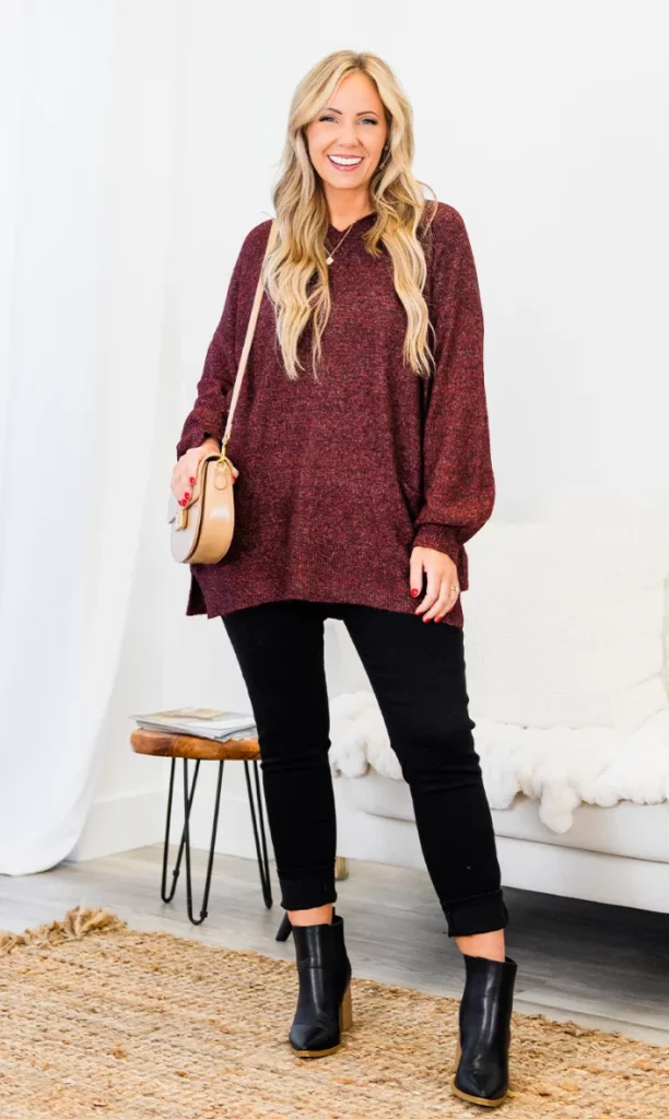 Woman in a loose burgundy outfit featuring a long sweater with black skinny pants, black ankle boots, and a beige crossbody bag.