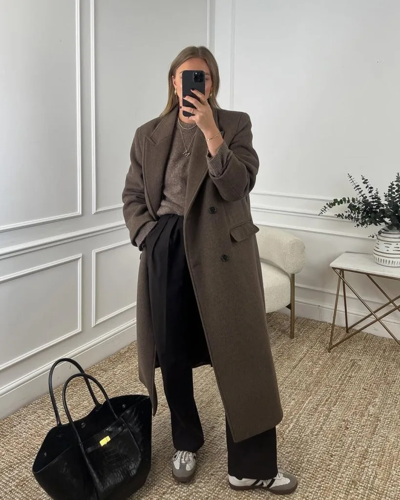 Office outfit in warm earthy tones – long brown wool coat over a matching sweater, paired with black tailored trousers, grey sneakers, and a large black textured tote bag.