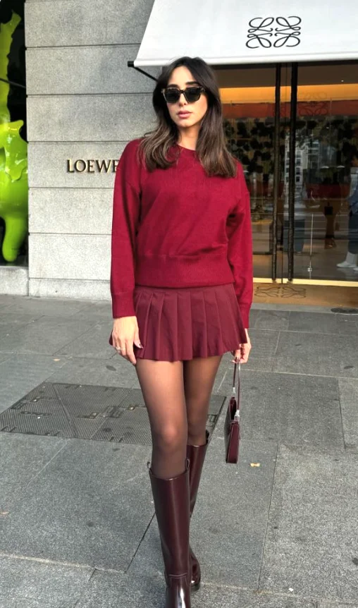 Woman wearing a chic burgundy outfit with a fitted sweater and matching pleated mini skirt, paired with sheer tights, burgundy knee-high boots, and a small matching handbag.