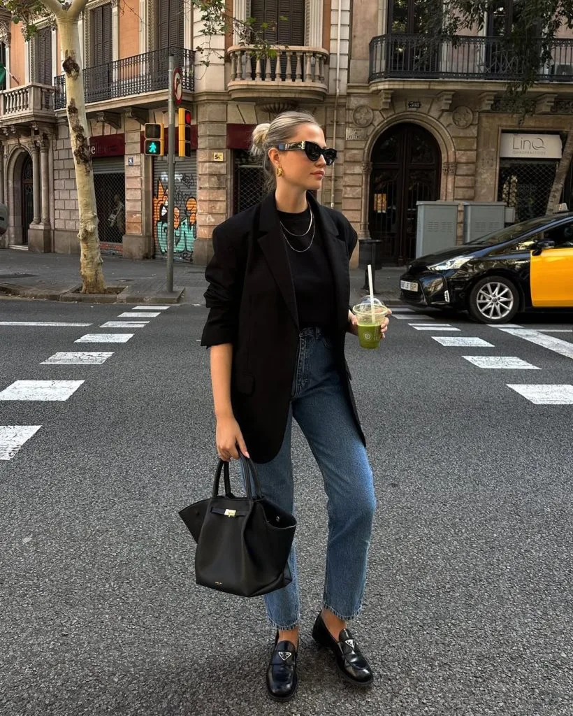 Office outfit with a modern edge – black blazer over a black T-shirt, blue straight-leg jeans, black loafers, and a structured black handbag.