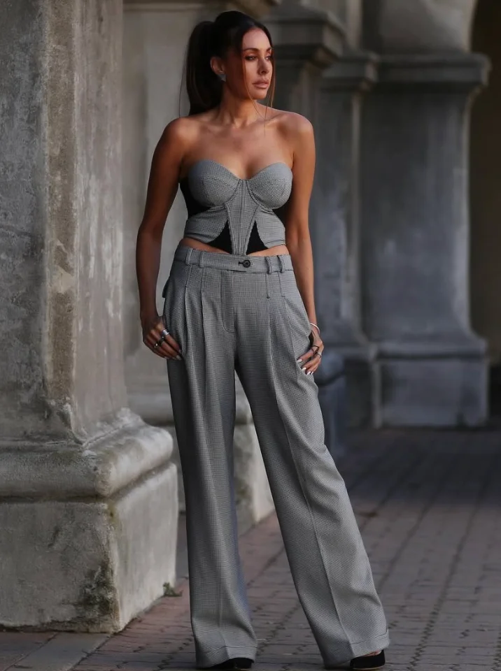 Statement grey trouser outfit featuring a strapless corset-style top with black cutouts, paired with wide-leg trousers and black heels, photographed in front of stone architecture.
