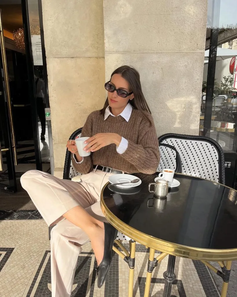 Office outfit with a classic Parisian touch – white collared shirt layered under a brown knit sweater, paired with cream trousers, black pointed flats, and oversized sunglasses.
