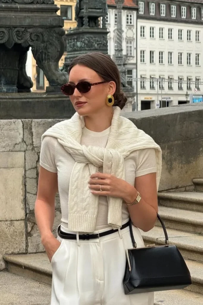 Old Money-Inspired Spring Outfit featuring a soft cream short-sleeve knit, tied sweater over the shoulders, and white pleated trousers with a black belt and bag.