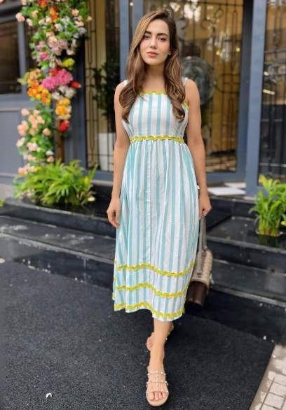 Woman in casual summer dress with vertical white and teal stripes, sleeveless with yellow trim details, paired with flat studded sandals.