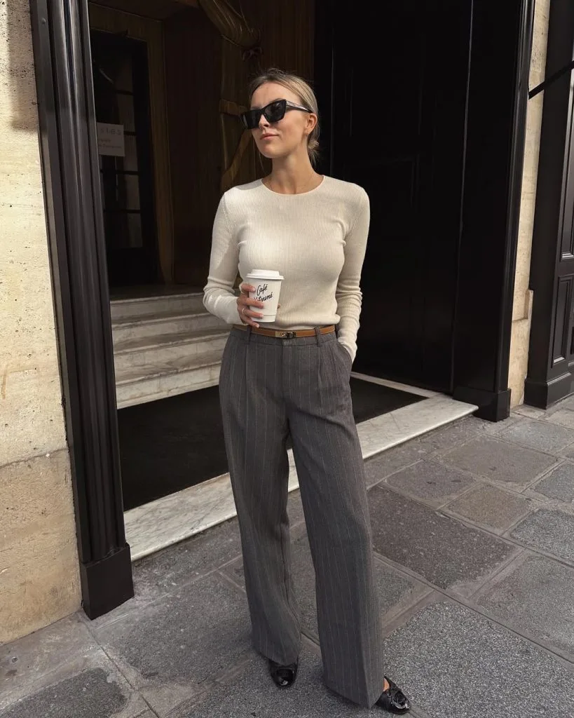 Office outfit in muted elegance – fitted cream ribbed long-sleeve top tucked into grey pinstripe wide-leg trousers, styled with black pointed-toe flats and dark sunglasses.