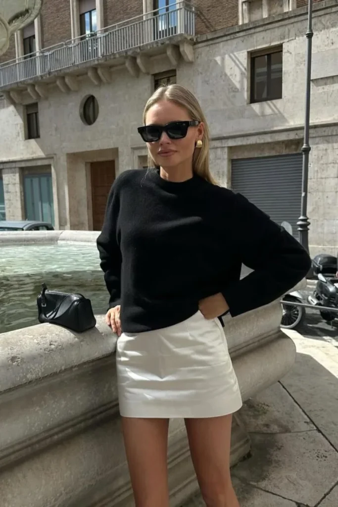 Old Money-Inspired Spring Outfit with a black sweater and white mini skirt, accessorized with gold earrings and a black handbag.