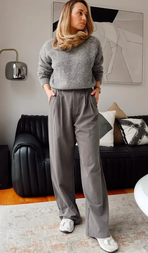 Relaxed grey trouser outfit with a soft grey sweater, loose pinstripe trousers, and white sneakers, photographed in a modern living room.