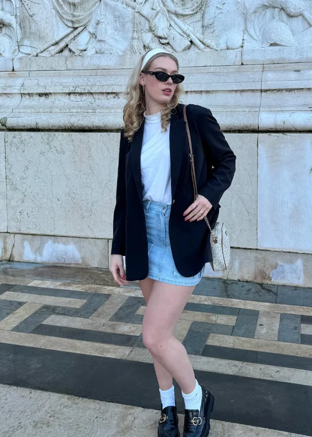 A cute simple outfit with a white t-shirt tucked into a light-wash denim mini skirt, layered with a black blazer and accessorized with loafers and a white headband for a classic preppy touch.