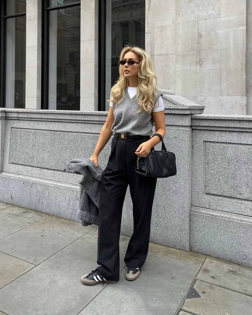 Office outfit with sporty sophistication – grey knit vest layered over a white T-shirt, paired with high-waisted black trousers, black Adidas sneakers, and a small black handbag.