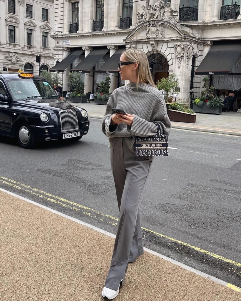 Woman in a grey trouser outfit styled with a chunky grey knit sweater, Christian Dior handbag, and white sneakers, standing on a city street.