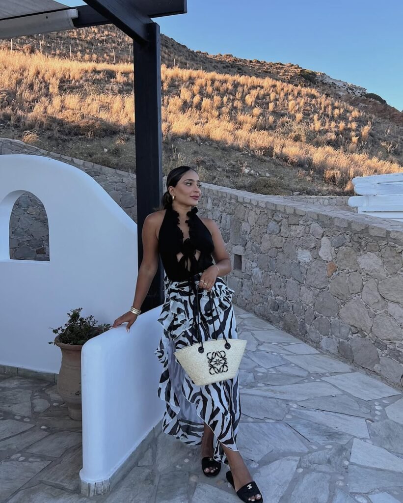 European Summer Outfit: Black sleeveless top with front tie detail paired with a zebra print ruffle skirt, black sandals, and a straw tote bag, posing on a terrace with a scenic hillside view.