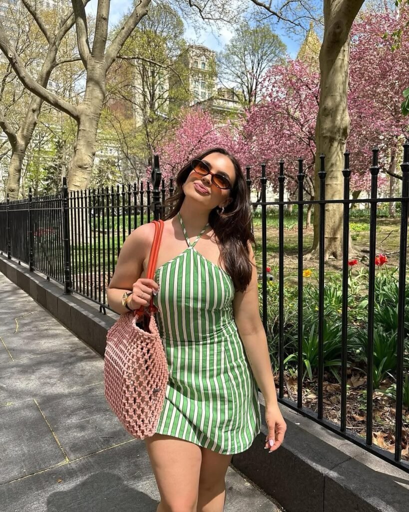 Woman in casual summer dress featuring green and white vertical stripes, halter neckline, paired with sunglasses, a pink woven tote bag, and walking in a sunny park with blooming trees.