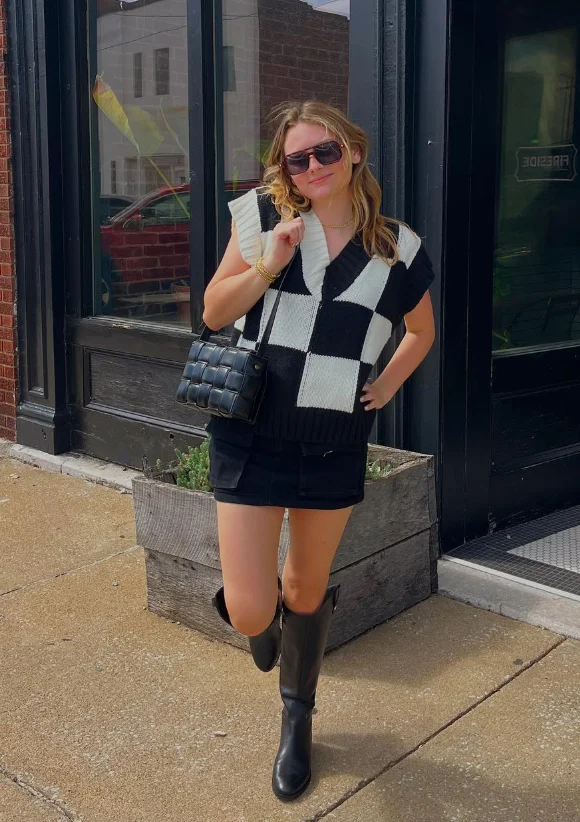 Early Fall Outfit with a black and white checkered sweater vest over a black mini skirt, paired with tall black boots and a quilted bag – cozy and chic for the season.