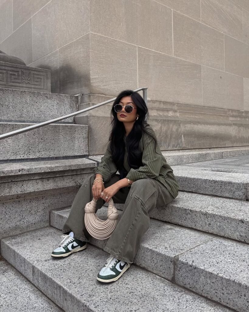 Woman in chill outfit ideas with an oversized green striped shirt, wide-leg cargo pants, green and white sneakers, and a unique circular beige bag sitting on stone steps.