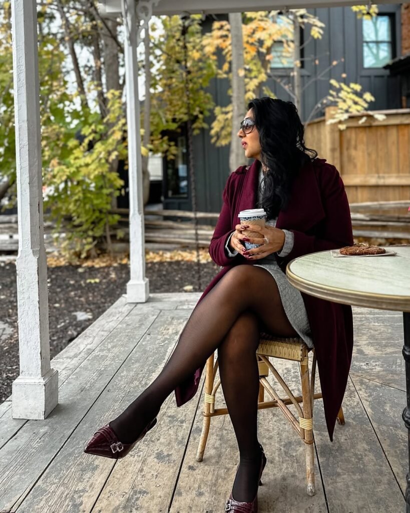 Woman in burgundy outfit with a long burgundy coat, grey dress, black tights, and burgundy heels, sitting at an outdoor café holding a coffee.