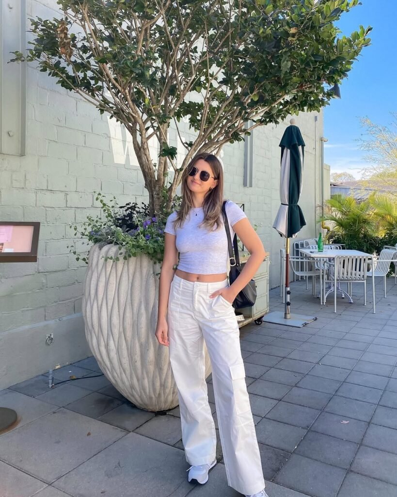 Woman in White Pants Outfits featuring a fitted light gray T-shirt, wide-leg white pants, and white sneakers, accessorized with sunglasses and a black bag.