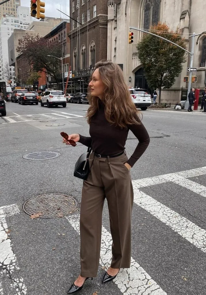 Office outfit in rich brown tones – fitted dark brown turtleneck tucked into high-waisted brown trousers, paired with black pointed heels and a small black crossbody bag.
