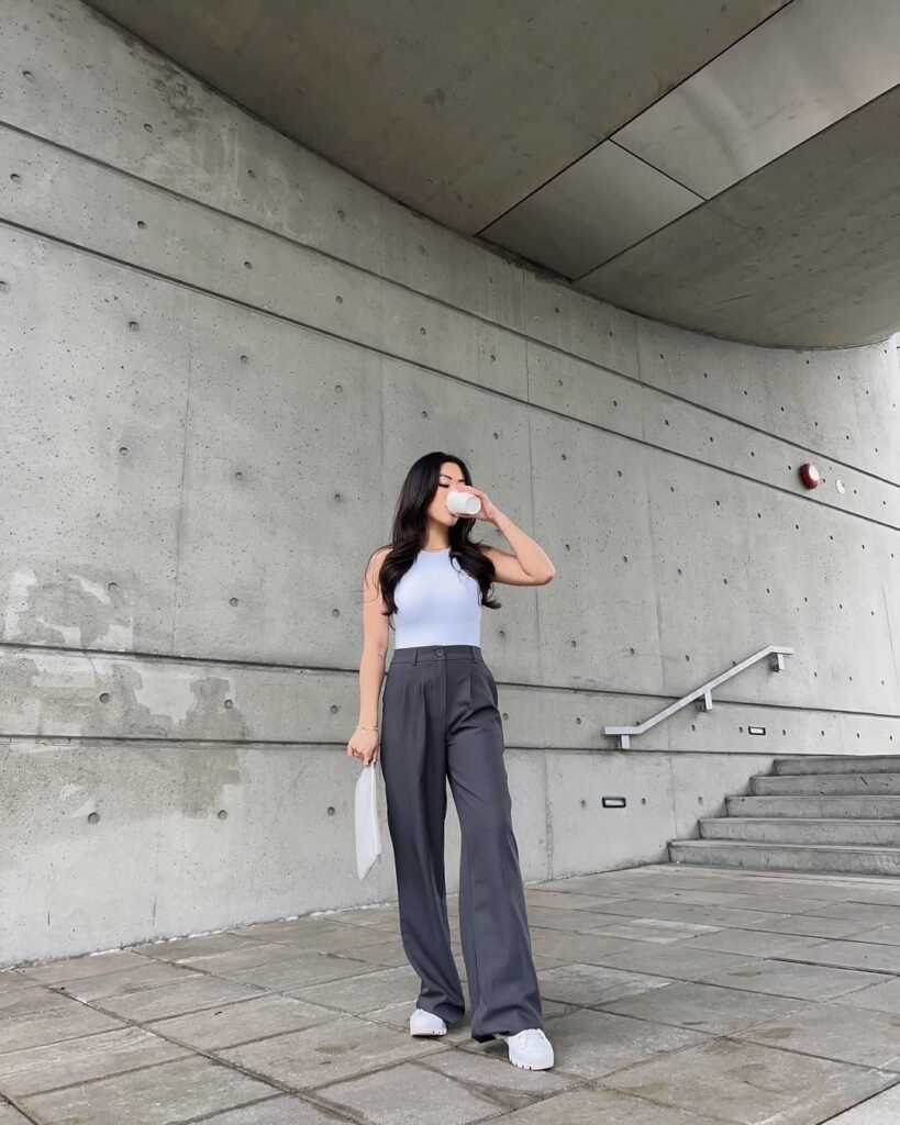 Minimalist grey trouser outfit featuring a white sleeveless top, white handbag, and white chunky shoes, photographed against a modern concrete wall.