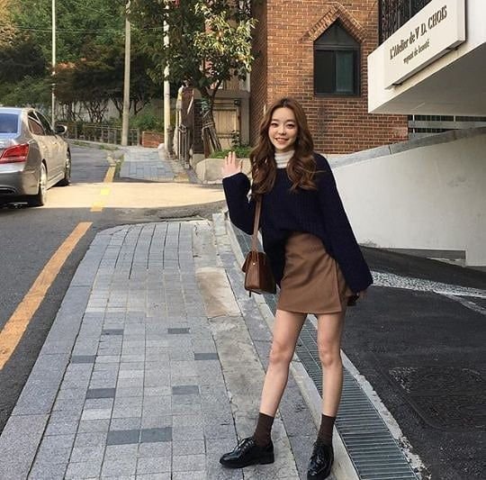 Woman in outfit korean style with a navy sweater, brown mini skirt, brown shoulder bag, and black shoes outdoors.