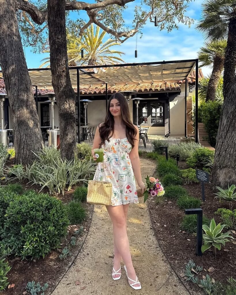 Woman wearing casual summer dress with a white base and colorful printed patterns, spaghetti straps, paired with white heels, holding flowers and a woven handbag in a garden.
