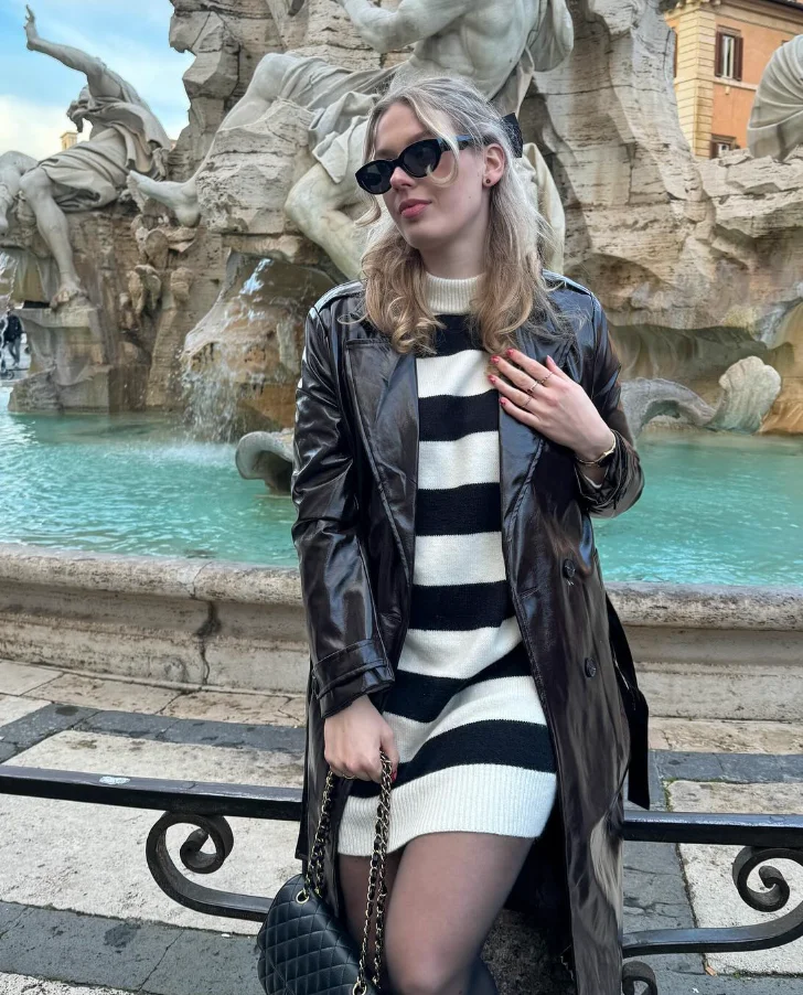 A woman sitting at a fountain in a black and white striped sweater dress layered with a black leather coat; a chic winter variation of cute simple outfits.
