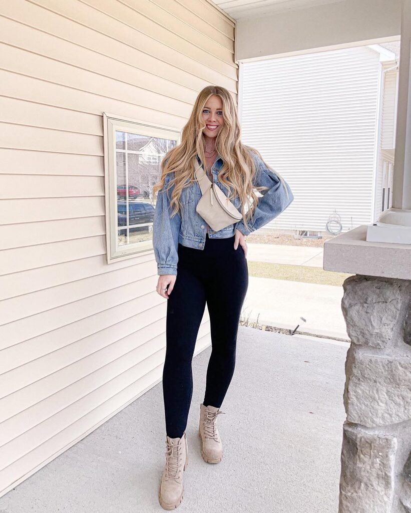 Woman in chill outfit ideas wearing a denim cropped jacket, black leggings, beige lace-up combat boots, and a cream crossbody bag, standing outdoors.