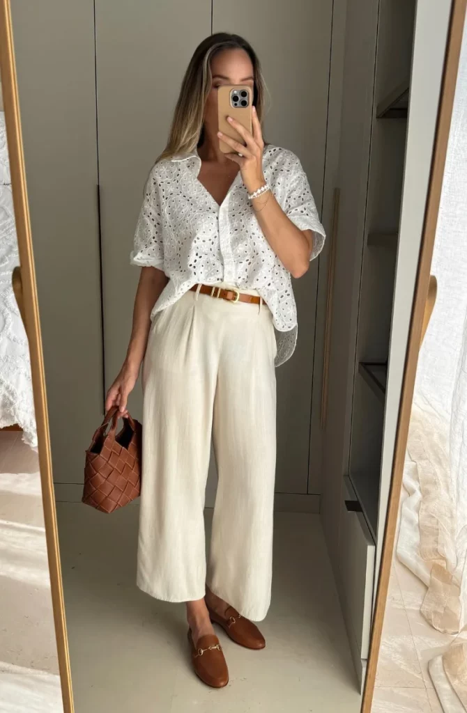 Office outfit in soft neutrals – a white embroidered short-sleeve blouse tucked into cropped cream trousers, styled with brown leather loafers, a woven handbag, and a matching belt.