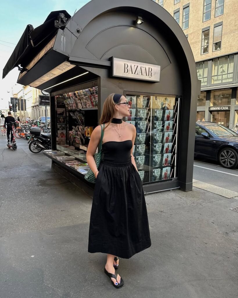 European Summer Outfit: Black strapless top with a matching black midi skirt, flip-flop sandals, and a green handbag, standing in front of a stylish newsstand.