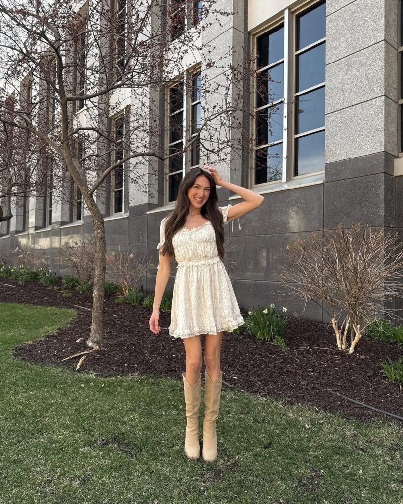 Woman in casual summer dress with a cream lace fabric, short puff sleeves, and a ruffled hem, paired with tan knee-high boots, posing outdoors near a building.