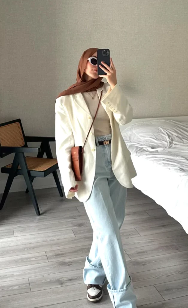 Office outfit with a relaxed twist – white oversized blazer over a beige turtleneck, light-wash wide-leg jeans, and brown-and-white sneakers, accessorized with a crossbody bag and sunglasses.