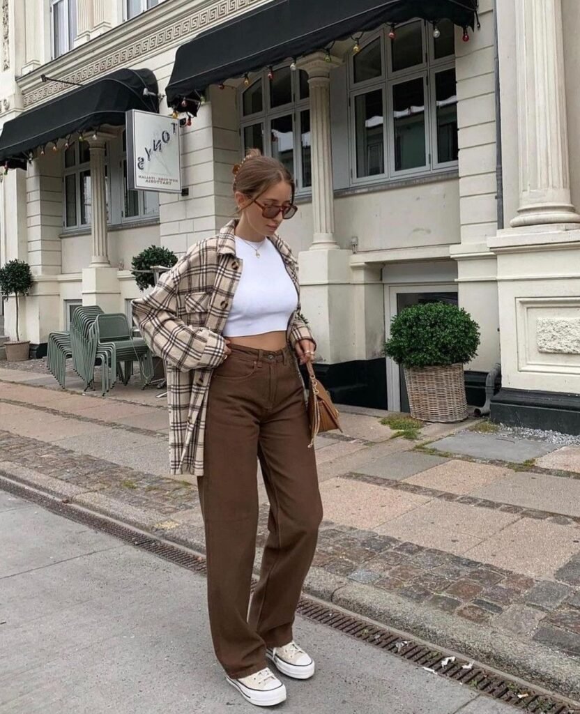 Brown Trousers Outfits – Woman wearing wide-leg brown trousers, a cropped white tank, and a beige plaid overshirt, paired with white platform sneakers and sunglasses.