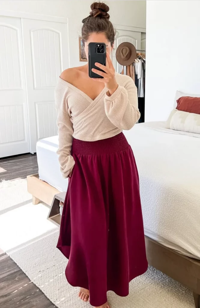 Woman in burgundy outfit wearing a beige off-shoulder sweater and a long burgundy skirt, posing indoors.