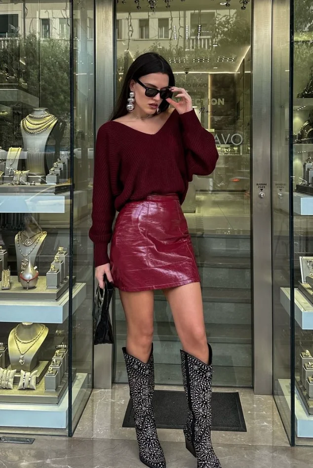 Woman in burgundy outfit with a burgundy sweater, burgundy leather mini skirt, and black embellished knee-high boots, posing outside a jewelry store.