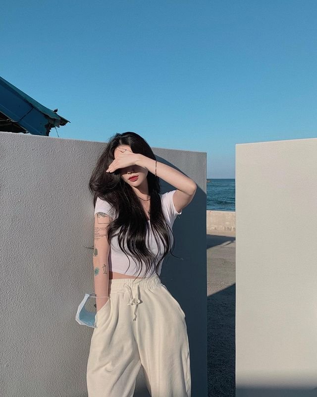 Woman in outfit korean style with a white fitted t-shirt and beige drawstring pants, posing outdoors by the sea.