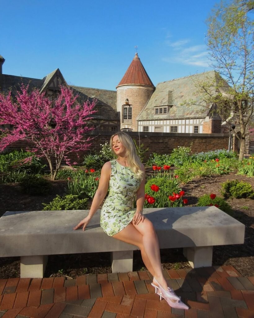 Woman wearing casual summer dress in light green with a floral print, sleeveless and fitted, paired with pastel pink heels, sitting on a stone bench in a blooming garden.