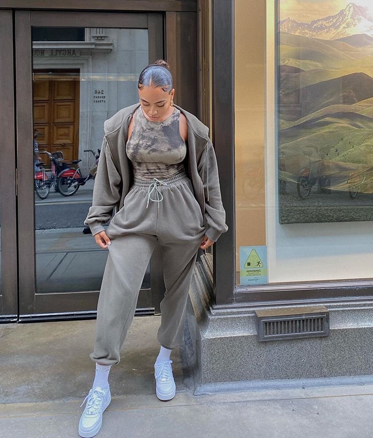 Woman in chill outfit ideas with a matching taupe tracksuit, tie-dye crop top, and white sneakers, standing on a city sidewalk.