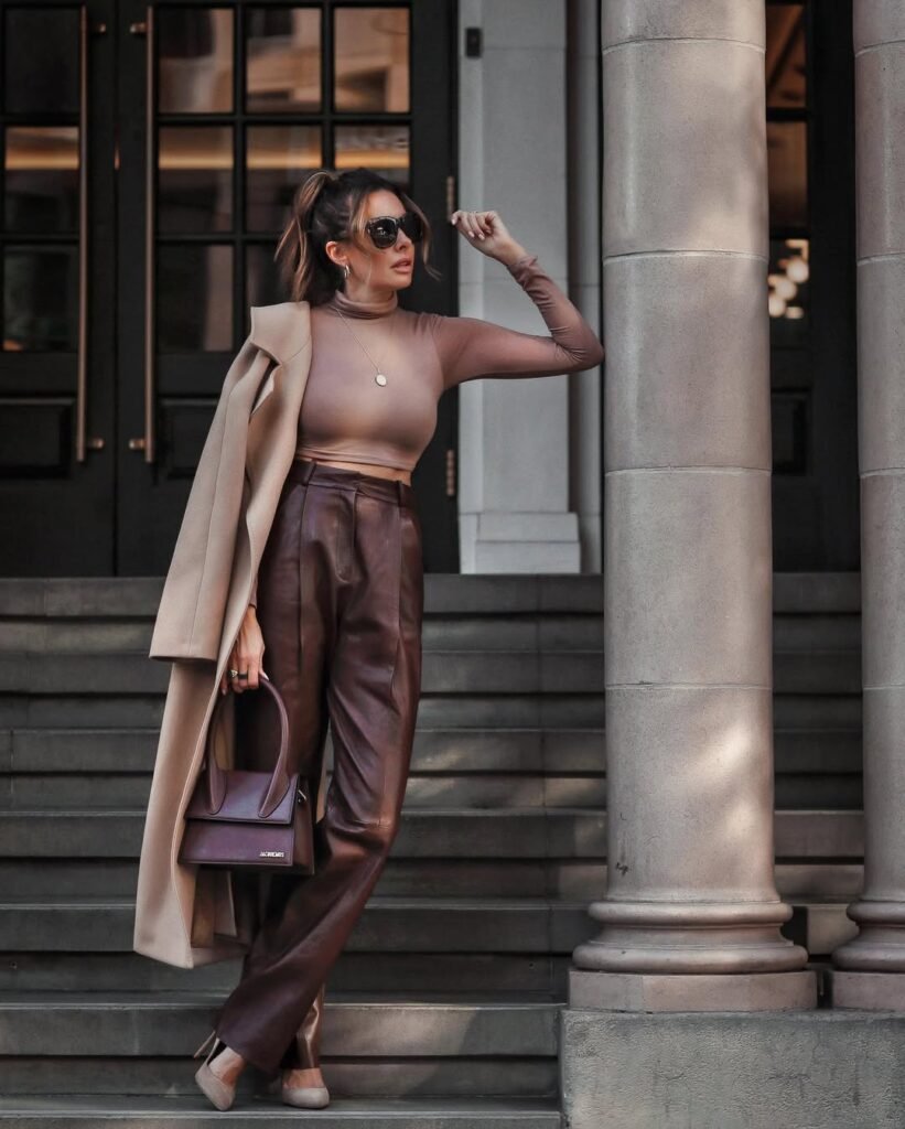 Brown Trousers Outfits – Woman standing on stone steps wearing brown leather trousers, a fitted brown long-sleeve top, beige overcoat draped over her shoulders, and matching beige heels, carrying a structured handbag.