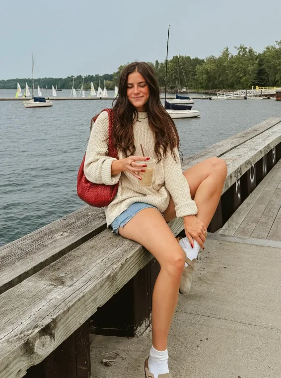 Early Fall Outfit with a cozy beige knit sweater layered over denim shorts, paired with white sneakers and a red shoulder bag – ideal for lakefront walks.