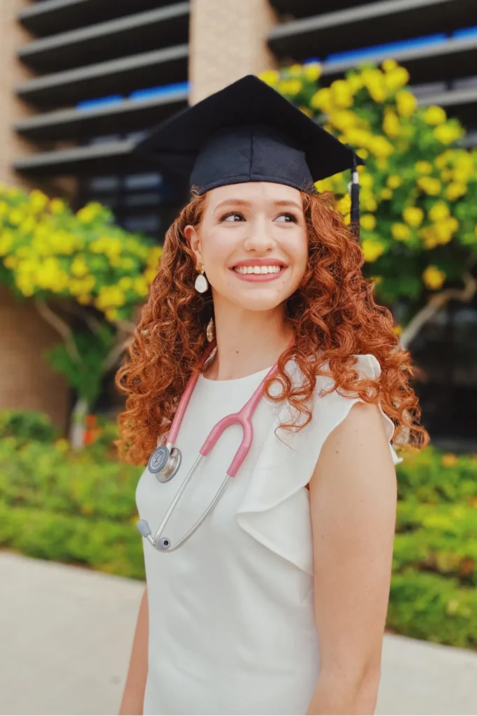 Graduation outfit ideas – elegant white dress with ruffle sleeves, paired with a graduation cap and pink stethoscope for a medical school graduation photoshoot.