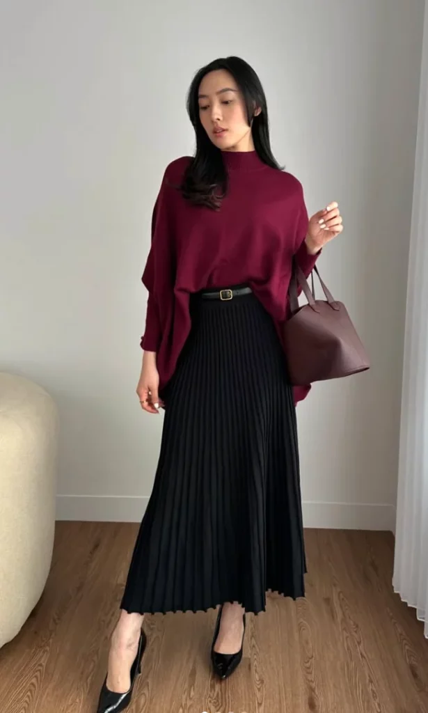 Woman in burgundy outfit wearing a burgundy poncho-style top, black pleated skirt, black heels, and carrying a burgundy handbag.