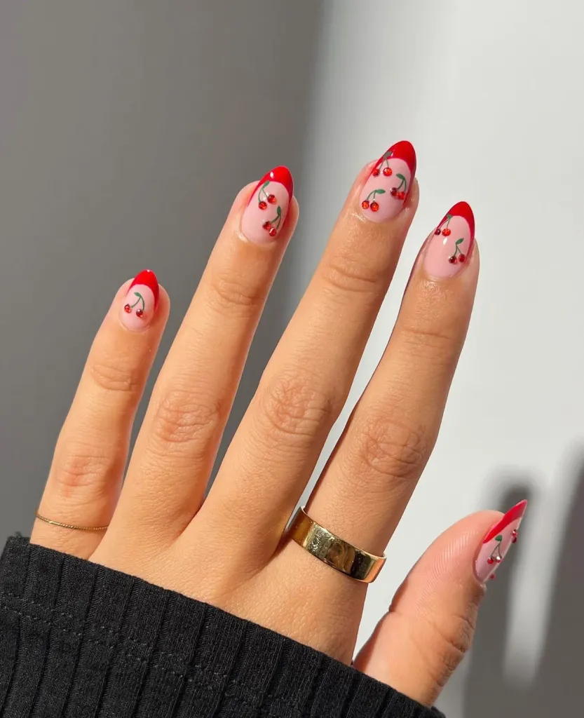 Almond nude nails with bold red French tips and scattered cherry nail art.