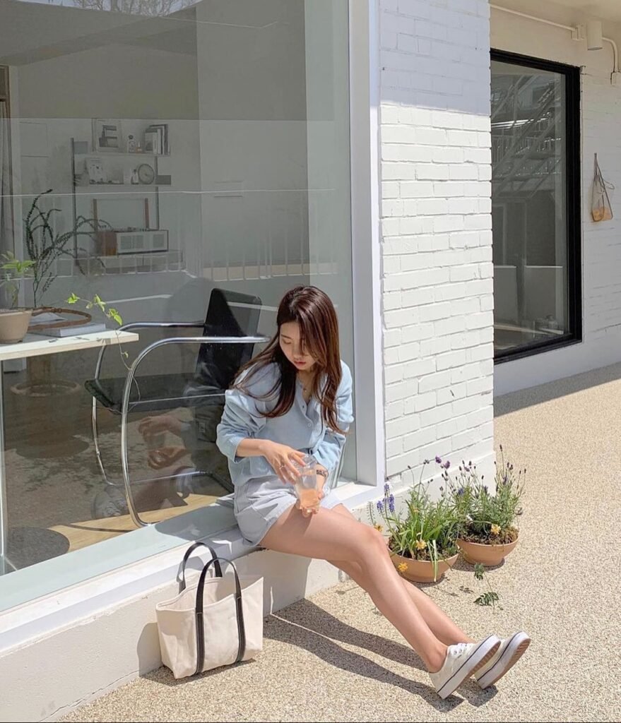 Woman in outfit korean style wearing a light blue blouse, beige mini skirt, and white sneakers sitting outside with a tote bag.