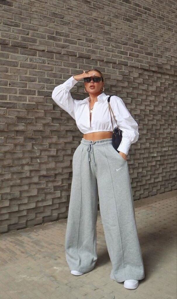 Woman in chill outfit ideas wearing a white cropped blouse, wide-leg grey sweatpants, white sneakers, and black sunglasses, holding a black shoulder bag.