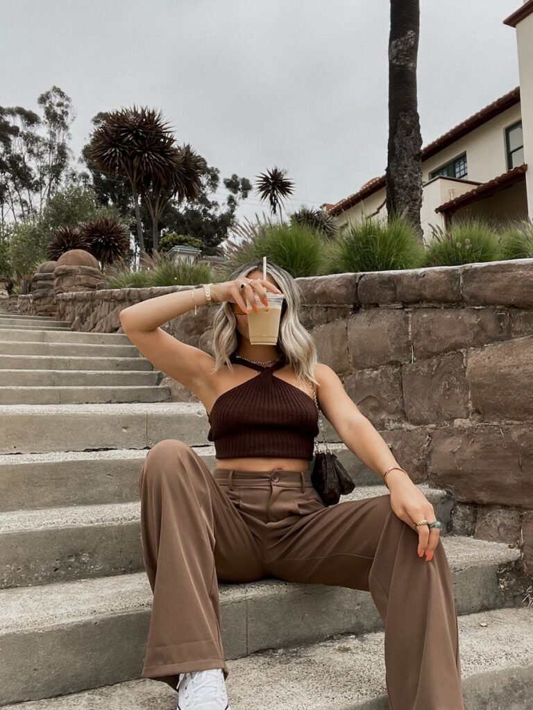 Brown Trousers Outfits – Woman sitting on outdoor stairs wearing light brown wide-leg trousers, a dark brown halter knit crop top, and white sneakers, holding an iced coffee in front of her face.