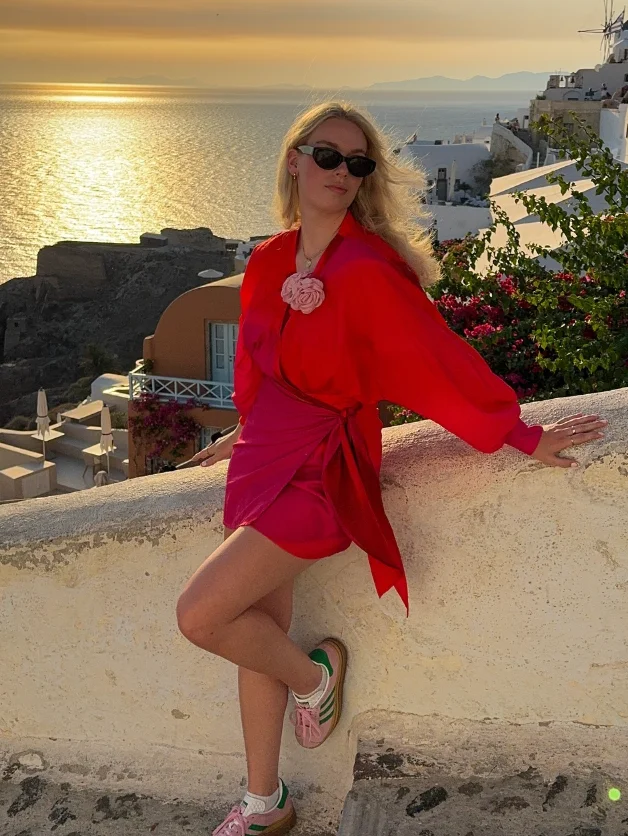 A woman leaning against a white wall at sunset in a bold red and pink wrap dress with pink sneakers, blending elegance with casual in a cute simple outfit.