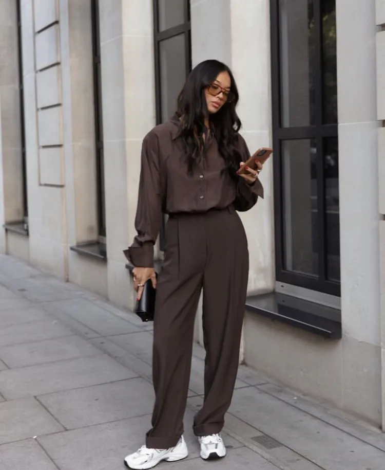 Brown Trousers Outfits – Woman in an all-brown relaxed fit ensemble with a button-up shirt and matching wide-leg trousers, paired with chunky white sneakers and a small clutch bag.
